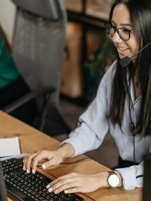 pexels photo 7658238 7658238 Cheerful woman in a call center typing on keyboard, wearing headset and eyeglasses.